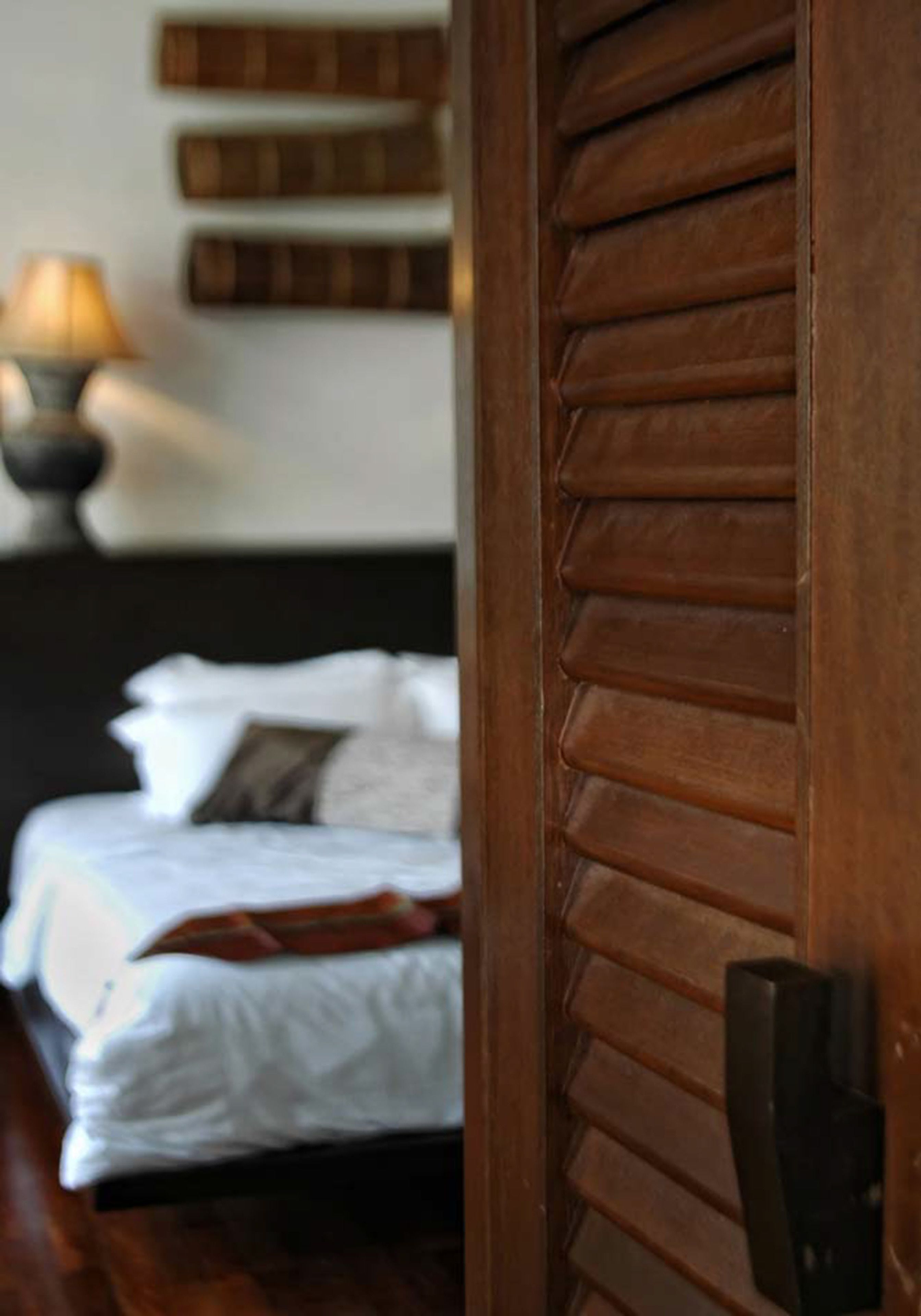 Una puerta de madera con listones horizontales, una cama con ropa de cama blanca y una almohada, y una lámpara fijada a la pared.