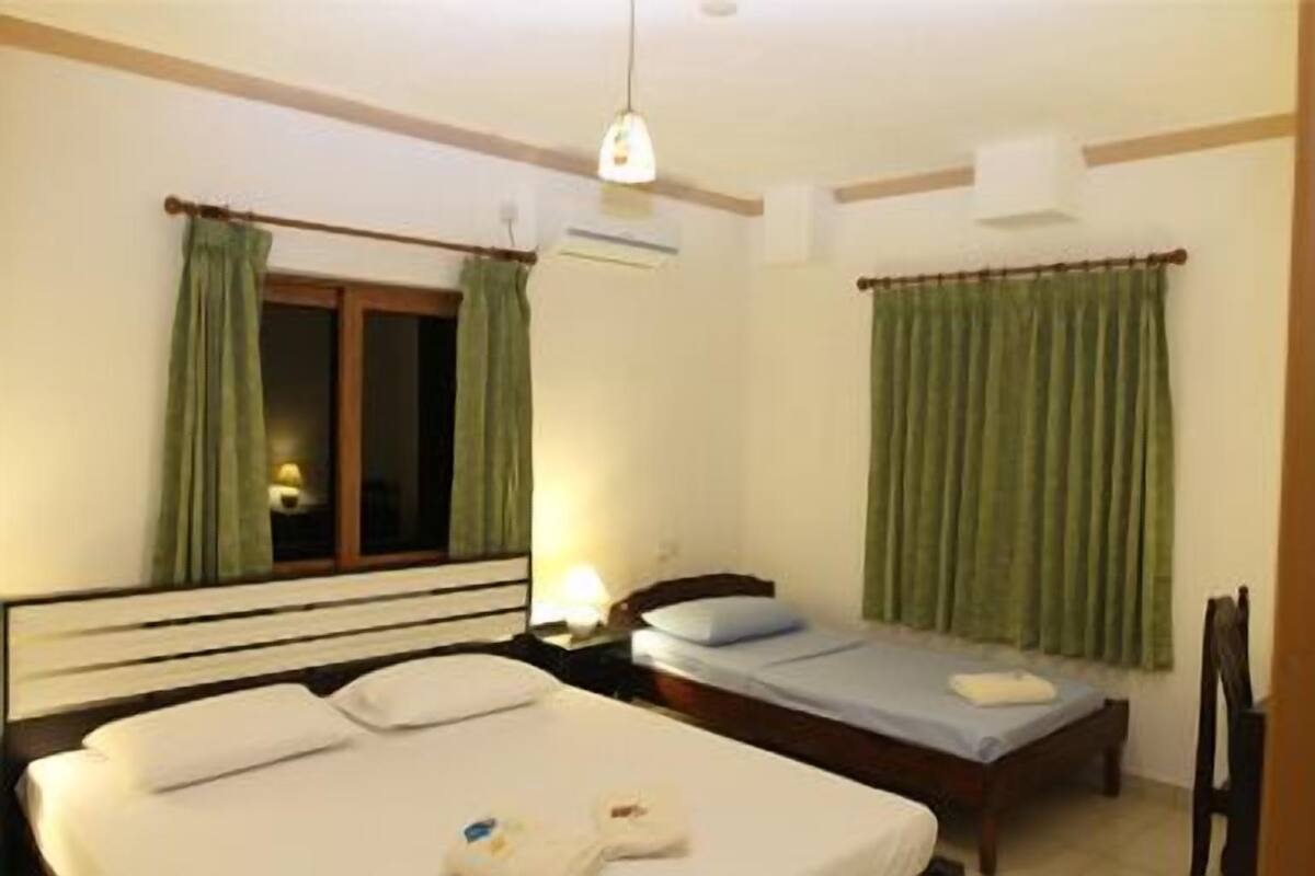 Ein Hotelzimmer mit einem großen Bett, einem Einzelbett, einem Nachttisch mit Lampe und einem Fenster mit Vorhängen.