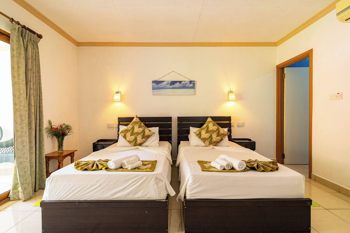 Ein Hotelzimmer mit zwei Betten, jeweils mit einem Kopfkissen und einem gerollten Handtuch. Es gibt einen kleinen Tisch mit einer Vase und einer Pflanze.