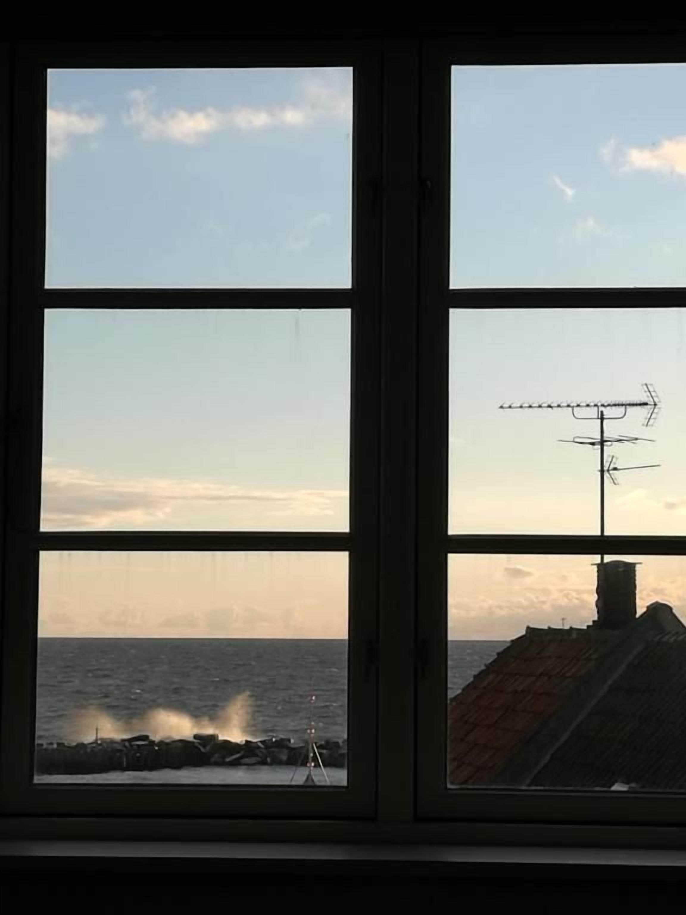 Et vindue med udsigt til havet og en bygning med skorsten.