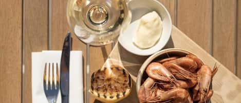 Een houten tafel met een schaal garnalen, een glas witte wijn, een kleine schaaltje boter en een bol knoflook.