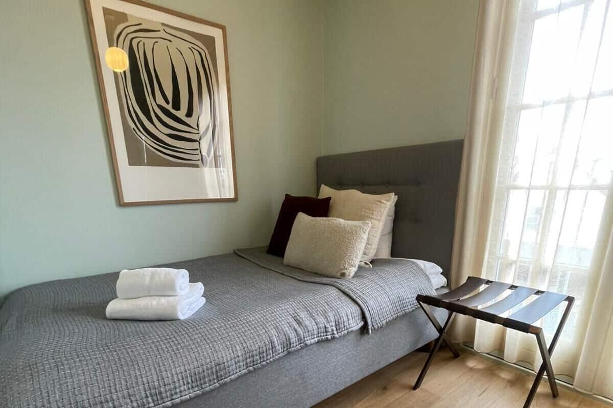 A single bed with a grey blanket, a headboard, and a bedside table with a lamp.