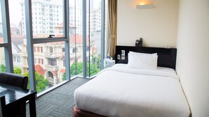 Quarto de hotel com uma cama grande, uma escrivaninha e vista para a paisagem urbana através de amplas janelas.