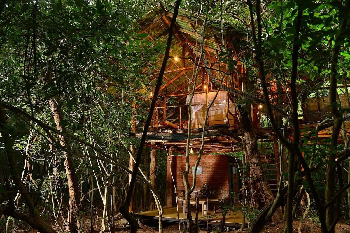 A rustic treehouse with a brick base and wooden upper levels, surrounded by dense forest.