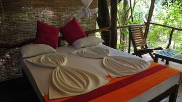 Una cama con almohadas ordenadas y una colcha a rayas, ubicada en una habitación con techo de madera y vistas al exterior lleno de vegetación.
