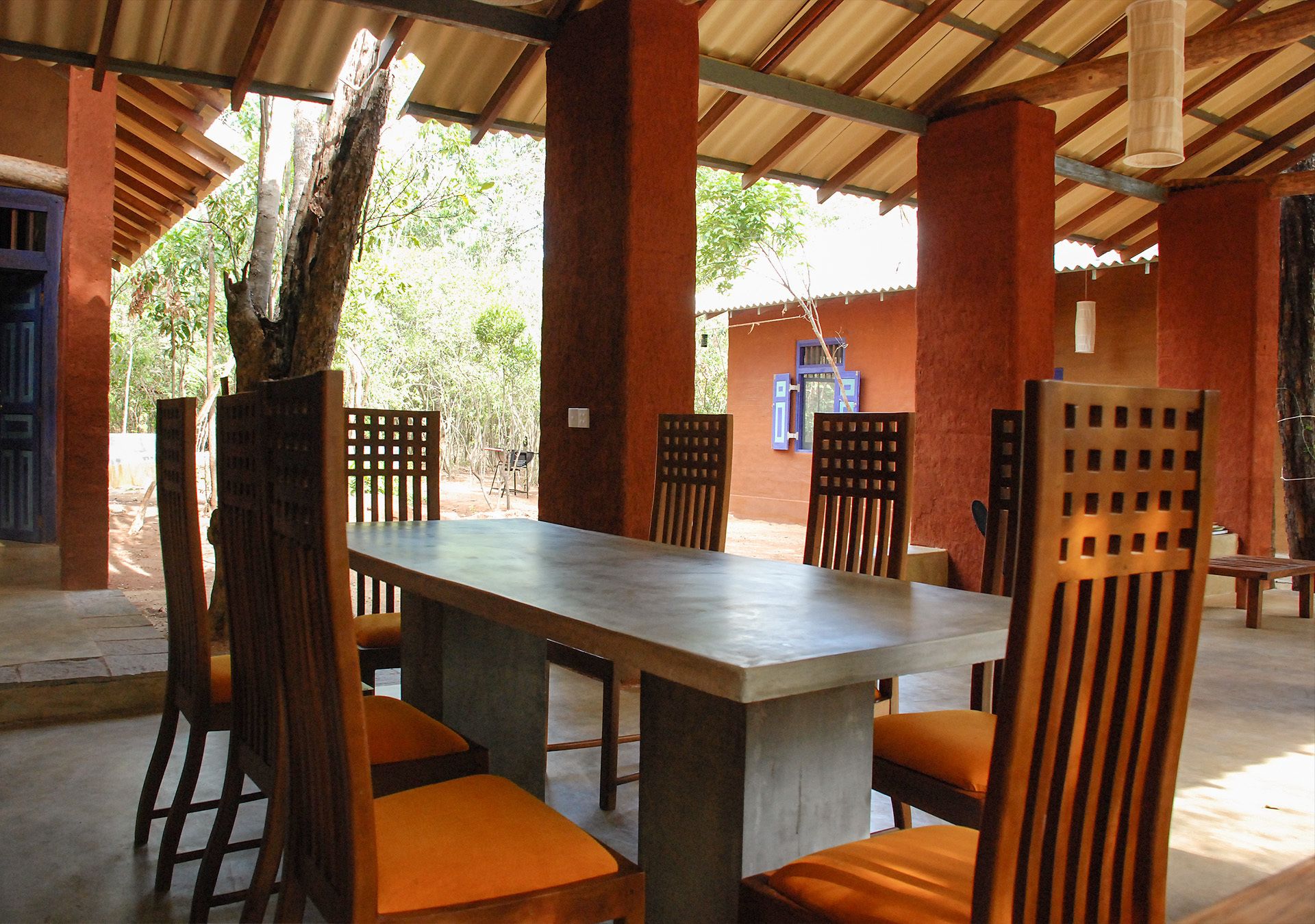 A covered outdoor dining area with a concrete table and wooden chairs with orange cushions.