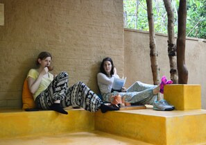 Two individuals seated on a yellow bench outdoors, one reading a book and the other holding a cup.