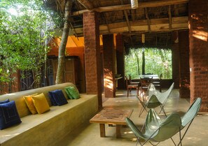 An outdoor seating area with a wooden bench and chairs, surrounded by trees and a thatched roof structure.