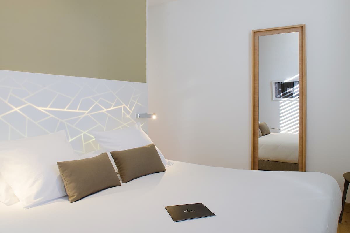 Cama bem arrumada com almofadas brancas e castanhas, um laptop na cama e um luminária geométrica na parede.