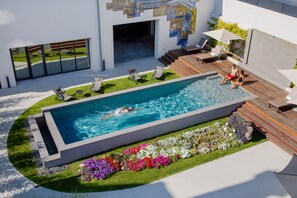 Une piscine extérieure moderne, dotée d’une terrasse en bois, de chaises longues et d’un jardin fleuri.