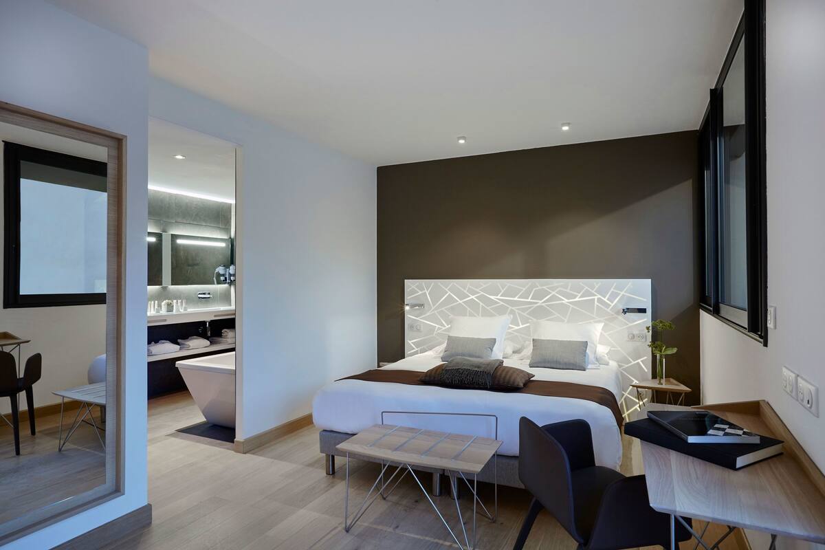 Quarto de hotel moderno com uma cama grande, uma escrivaninha, uma cadeira, uma mesinha e um banheiro com banheira.