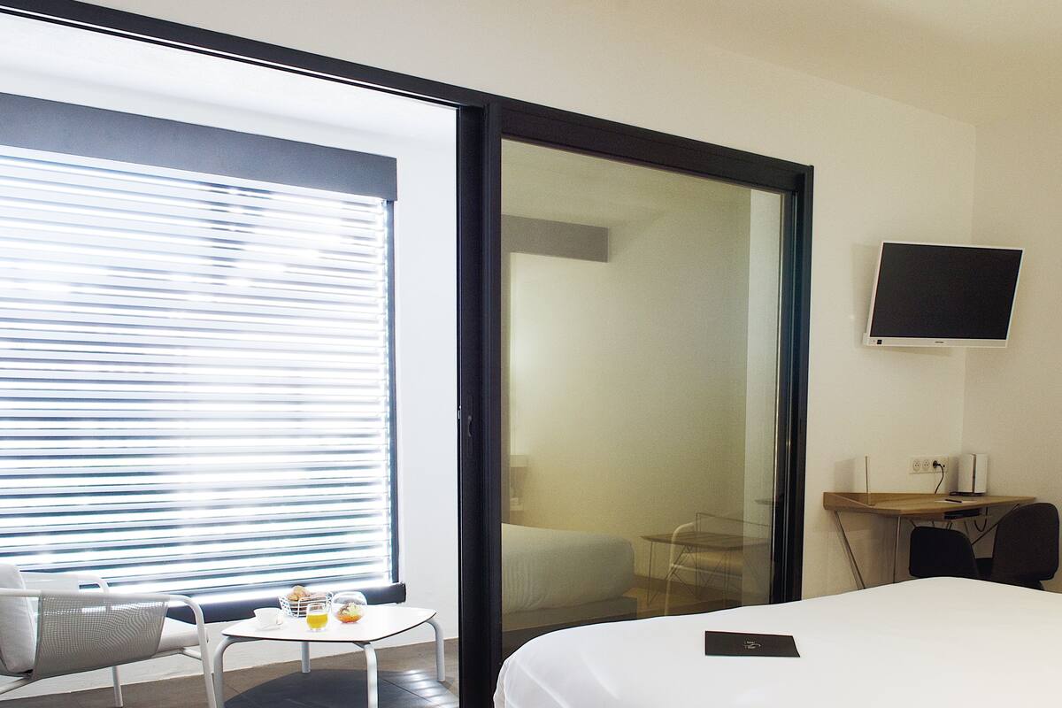 Quarto de hotel com cama, escrivaninha com computador, televisão, janela com persianas e pequena mesa com itens de café da manhã.