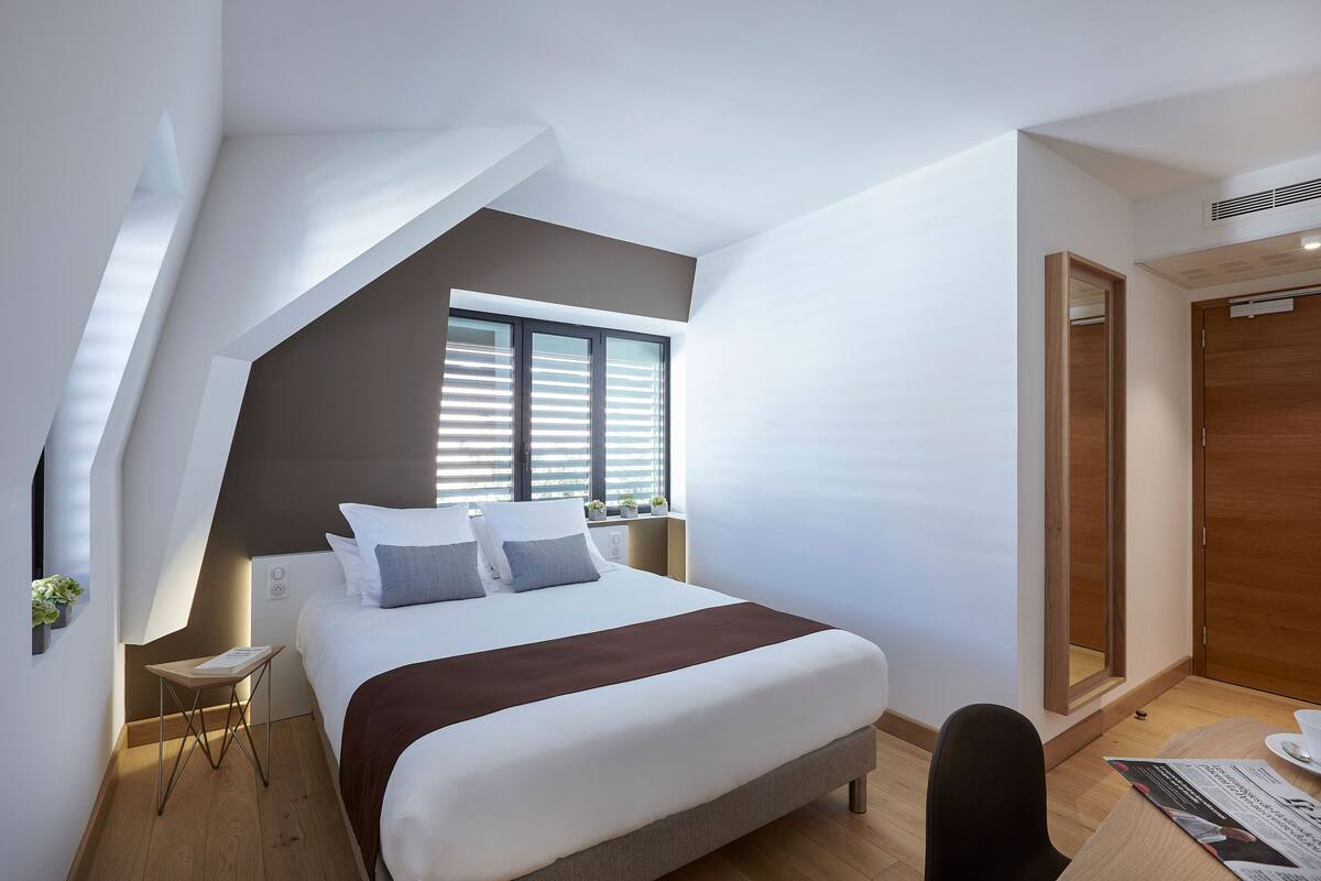 Um quarto moderno com uma cama grande, uma mesinha, uma cadeira e uma janela com persianas.