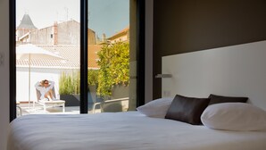 A hotel room with a large bed, a view of a balcony with a person sitting, and a cityscape in the background. - Hôtel In Situ (Béziers)