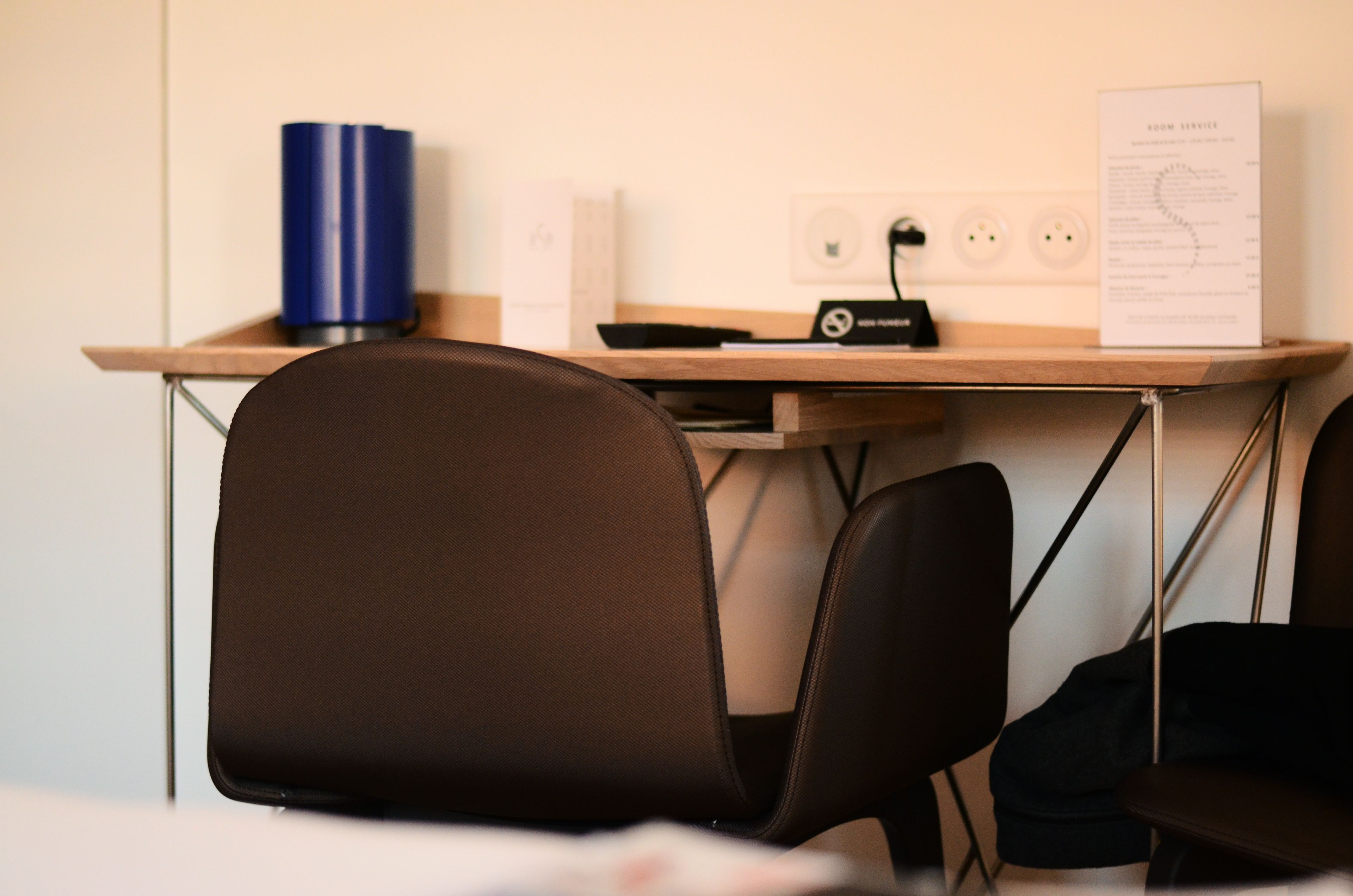 A hotel room with a desk, a blue cylindrical object, a black electronic device, and a menu on the wall.