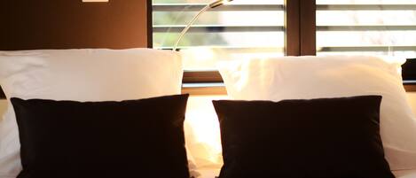 Habitación de hotel con una cama, almohadas blancas y negras, una mesita de noche y una ventana con persianas.