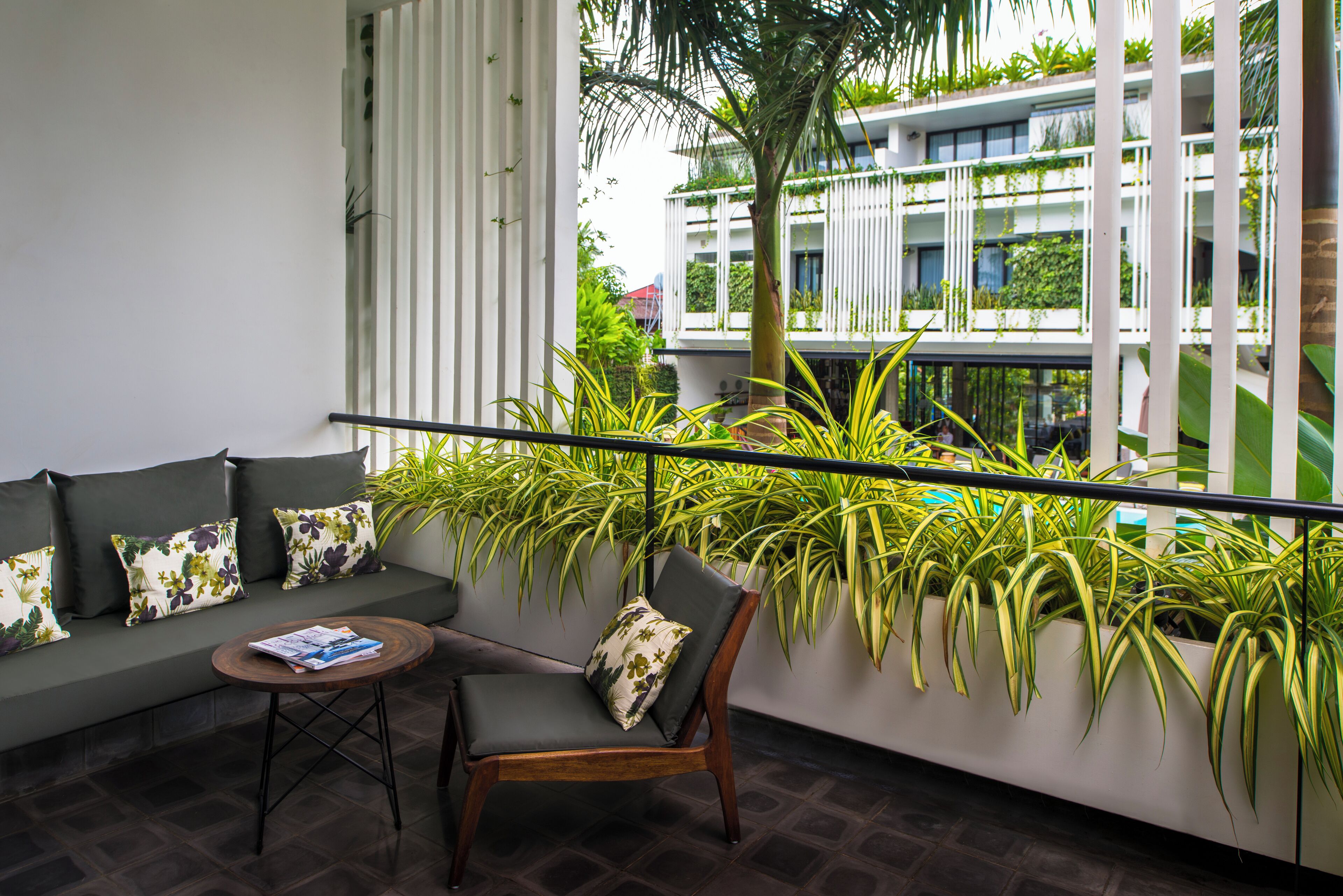 Un balcón con sofá y sillón, plantas en macetas y vista a un edificio con balcones.