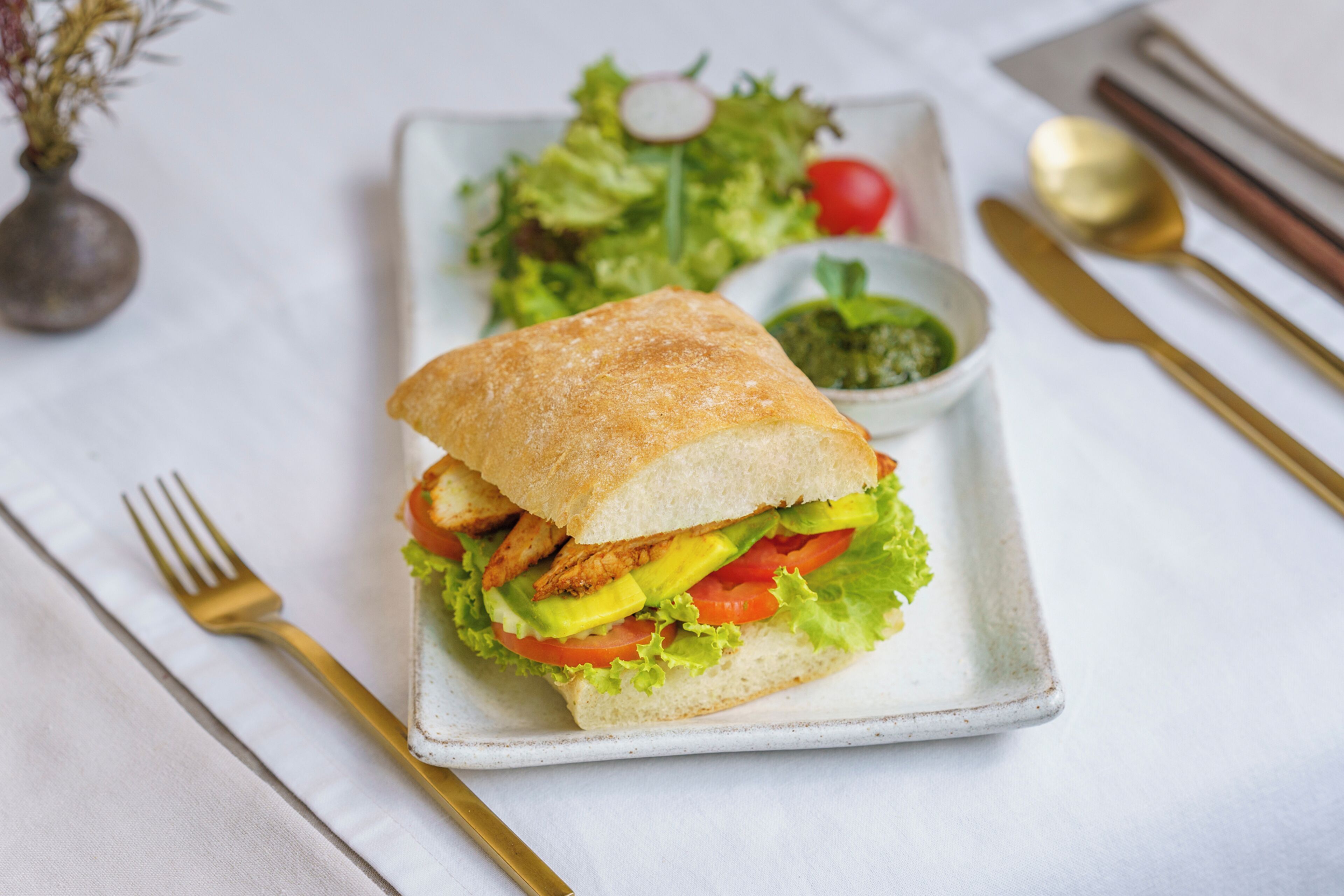 A sandwich on a white plate with a side salad and a small bowl of sauce.