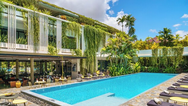 Une piscine entourée de verdure et dotée d’une terrasse en pierre, avec des chaises longues et des tables.