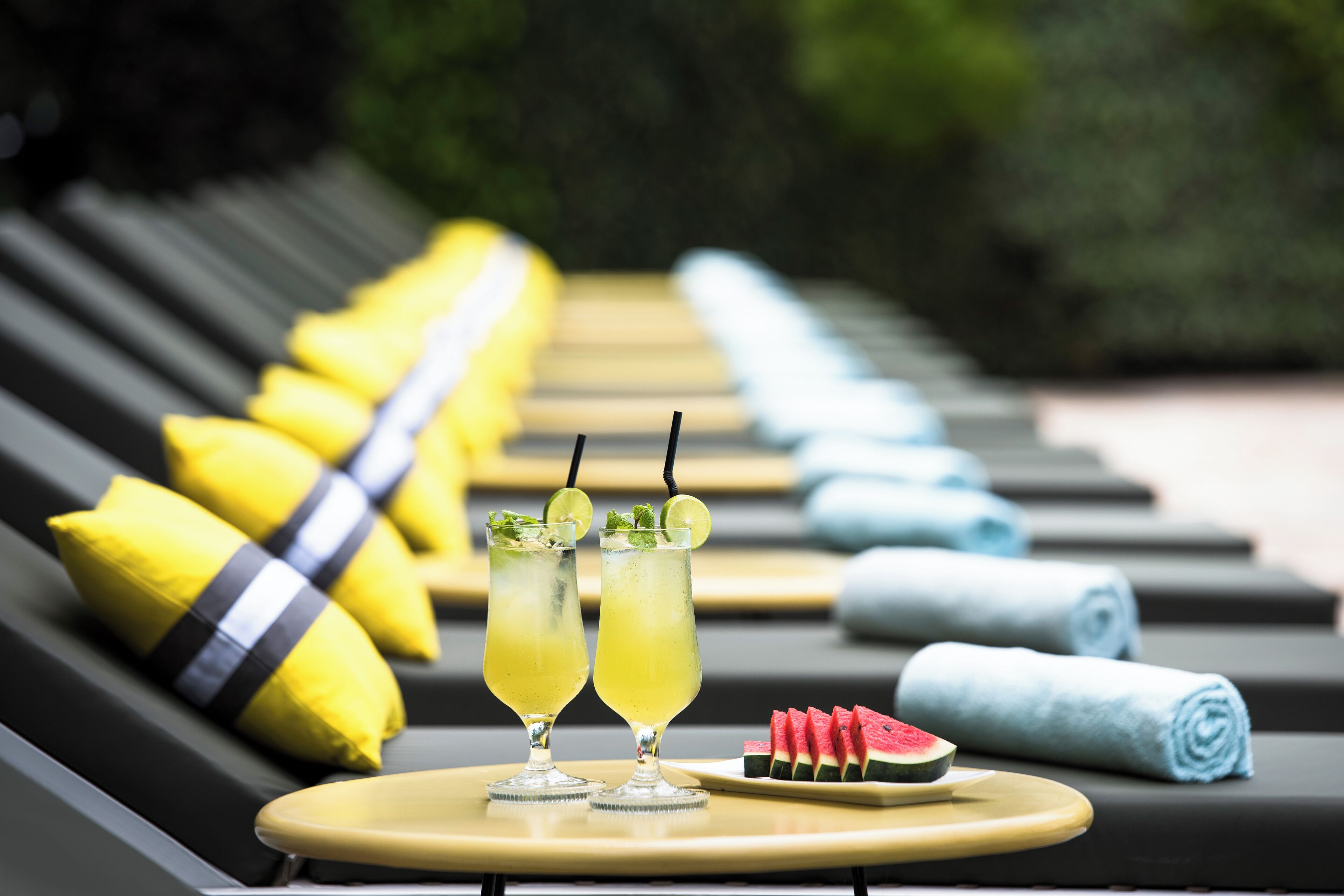 Twee cocktails op een dienblad met een stukje watermeloen, gele en blauwe handdoeken en gele kussens op lounge stoelen.
