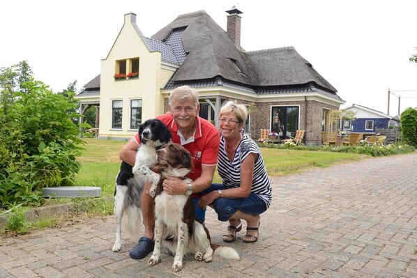 A family with two dogs in front of a house with a steeply pitched roof and a brick chimney. - Restinn Oosterleek (Oosterleek)