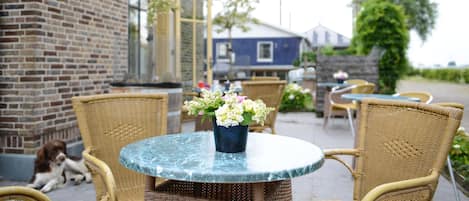 A round table with a vase of flowers, wicker chairs, and a brick building in the background.