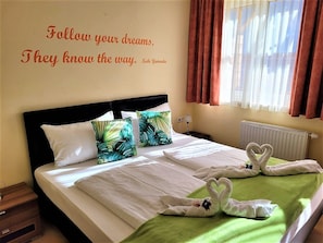 A neatly made bed with white linens, a green throw blanket, and decorative swan-shaped towels. A motivational quote is displayed on the wall.