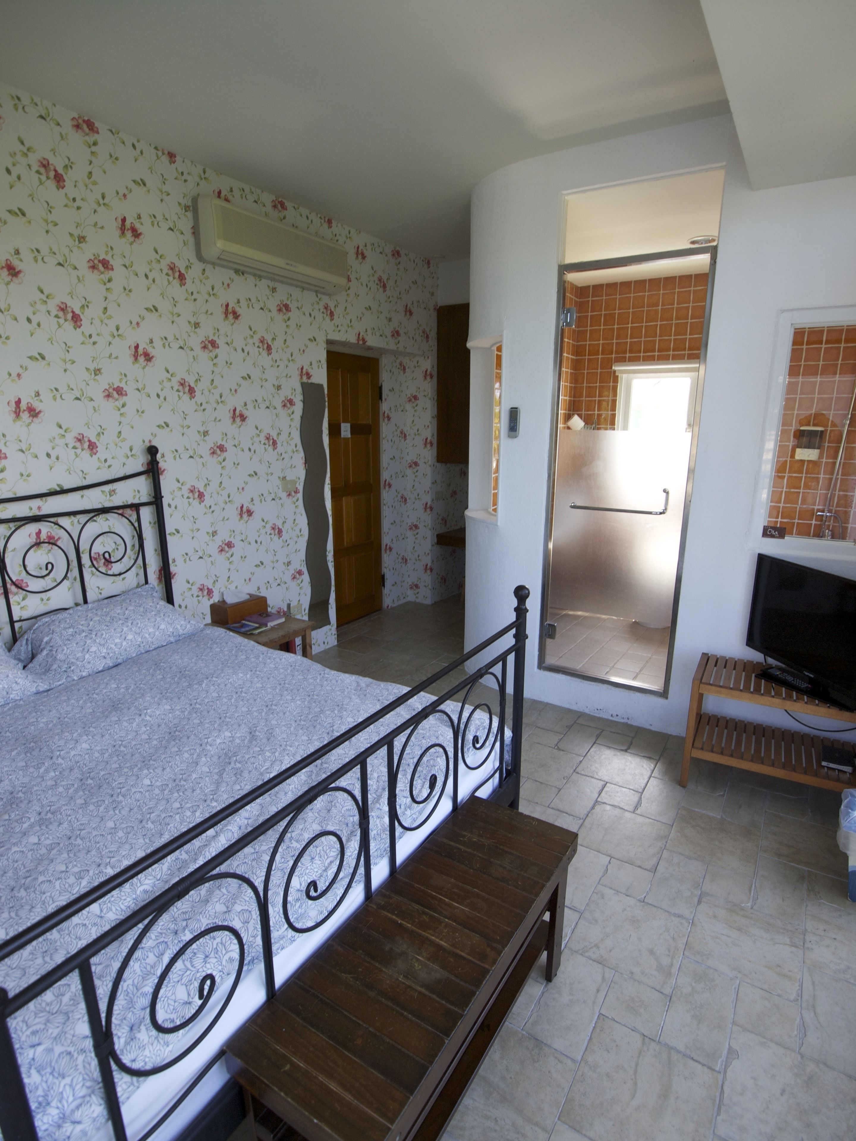Une chambre à coucher avec un lit, un banc en bois, un téléviseur et une salle de bain visible à travers une porte ouverte.