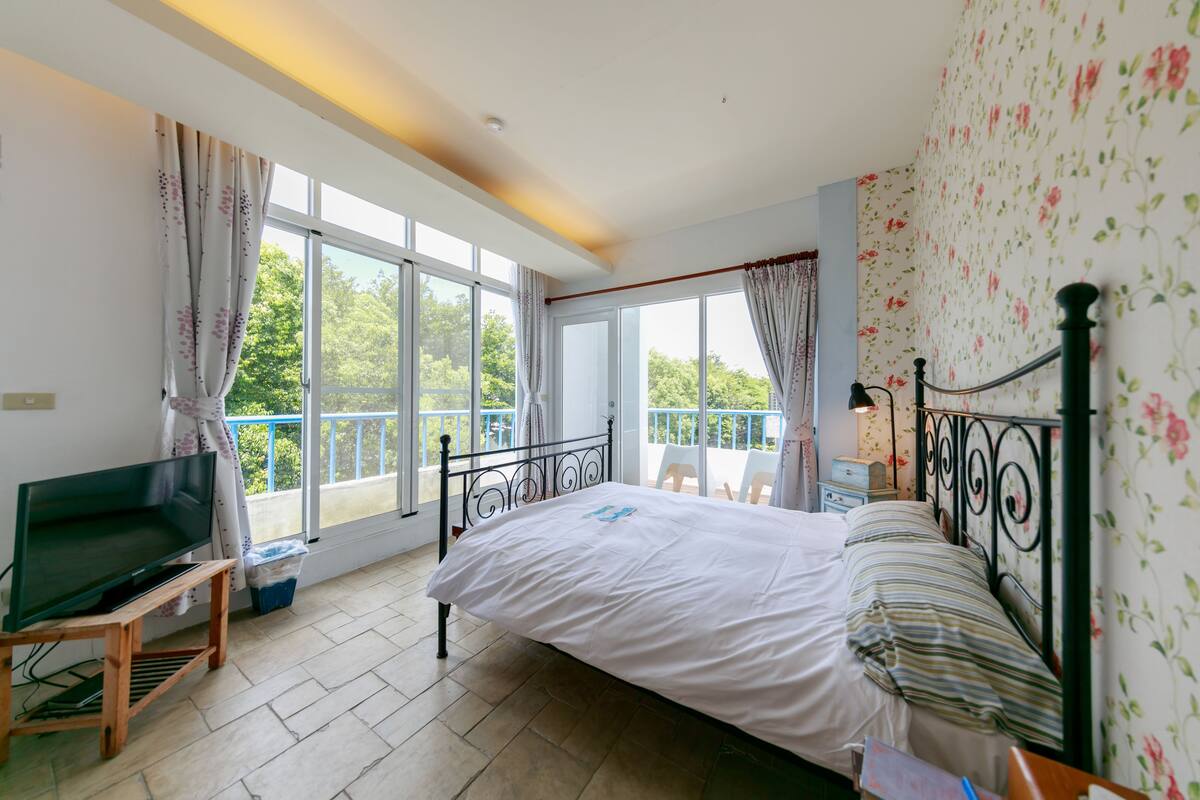 Un dormitorio con cama, televisor sobre un mueble, balcón con vistas a árboles y ventana con cortinas.