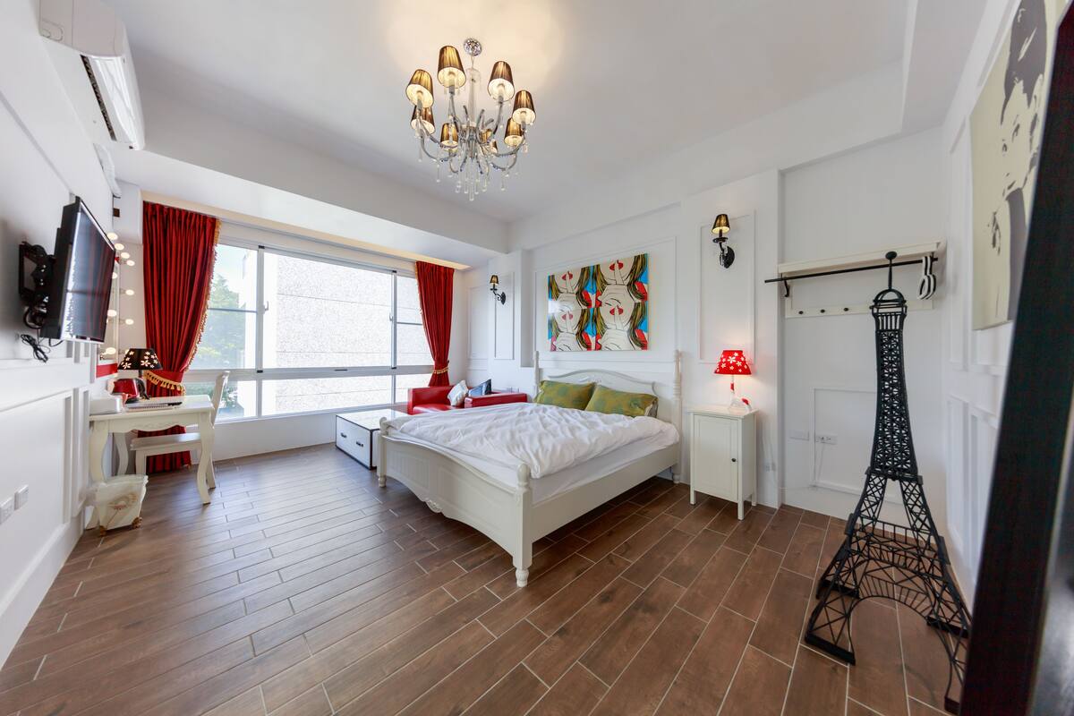 Un dormitorio con cama, escritorio, silla, televisor, ventana con cortinas, candelabro, obra de arte en la pared, mesita de noche, lámpara y una reproducción decorativa de la Torre Eiffel.