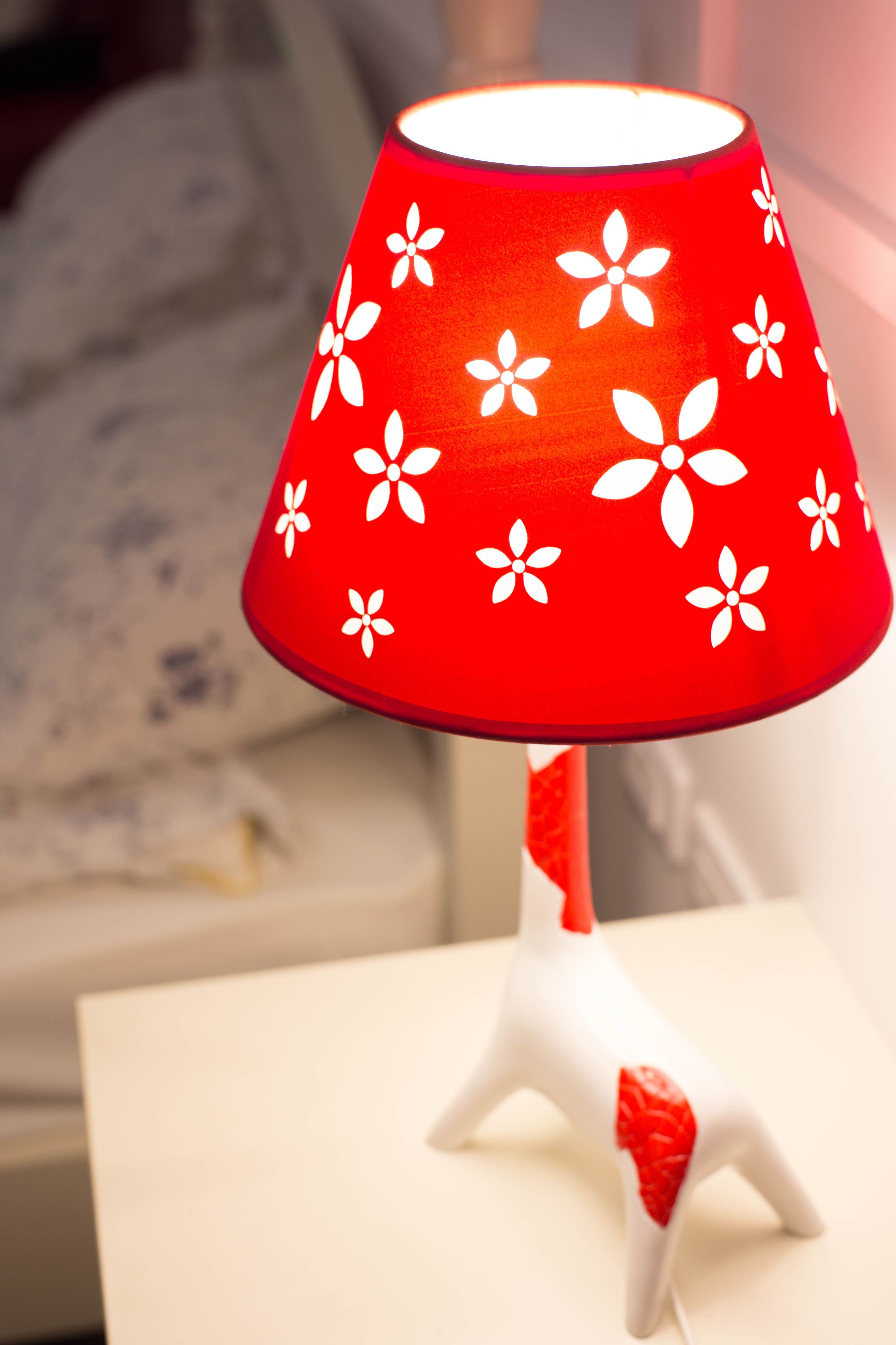 Luminária com cúpula vermelha estampada com motivos florais brancos, sobre superfície branca.