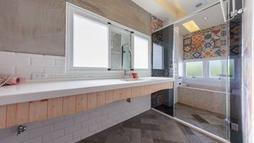 A modern bathroom with a white countertop, wooden paneling, a large mirror, a shower enclosure, and patterned tiles on the walls.
