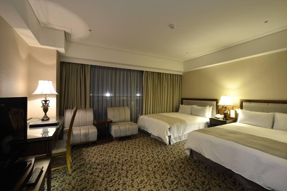A hotel room with two beds, a desk with a lamp, a chair, and a television.