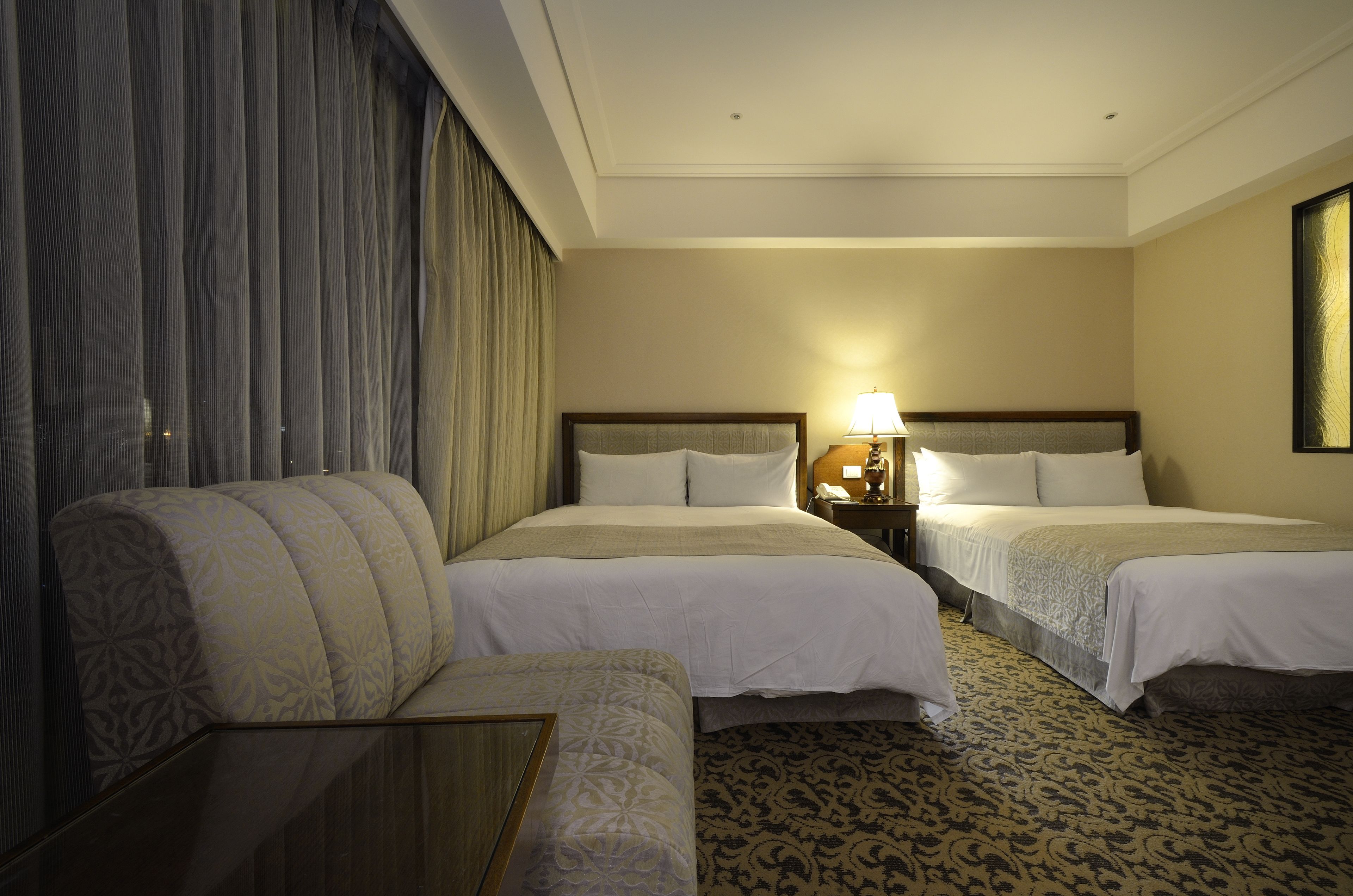 Habitación de hotel con dos camas, un sofá, una mesita de noche con lámpara y una alfombra estampada.