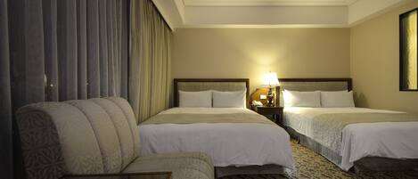 Habitación de hotel con dos camas, un sofá, una mesita de noche con lámpara y una alfombra estampada.