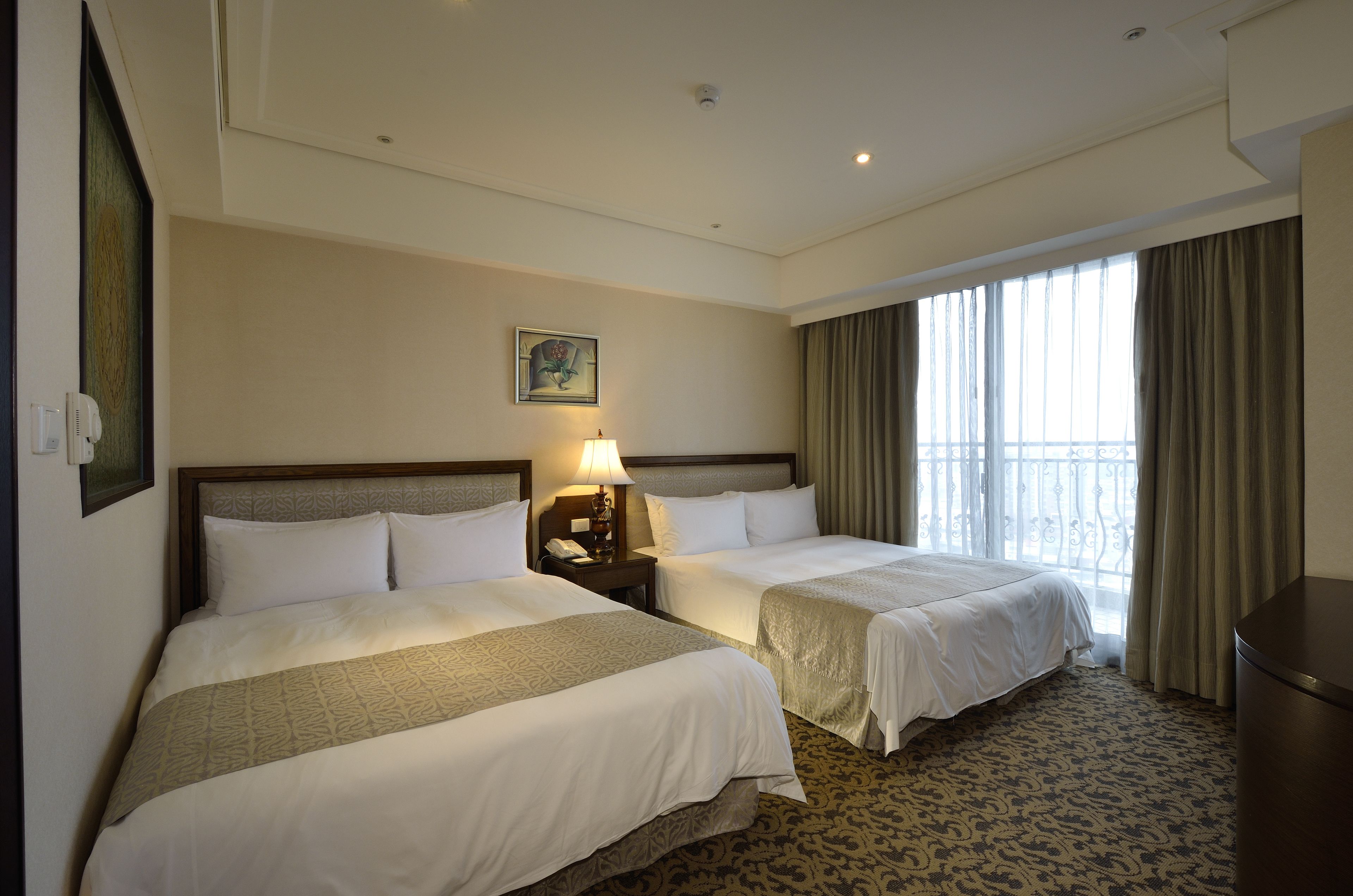 A hotel room with two beds, a nightstand with a lamp, a painting on the wall, and a large window with curtains.