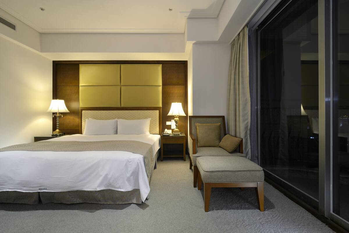 A hotel room with a large bed, two bedside tables with lamps, a chair, and a large window.