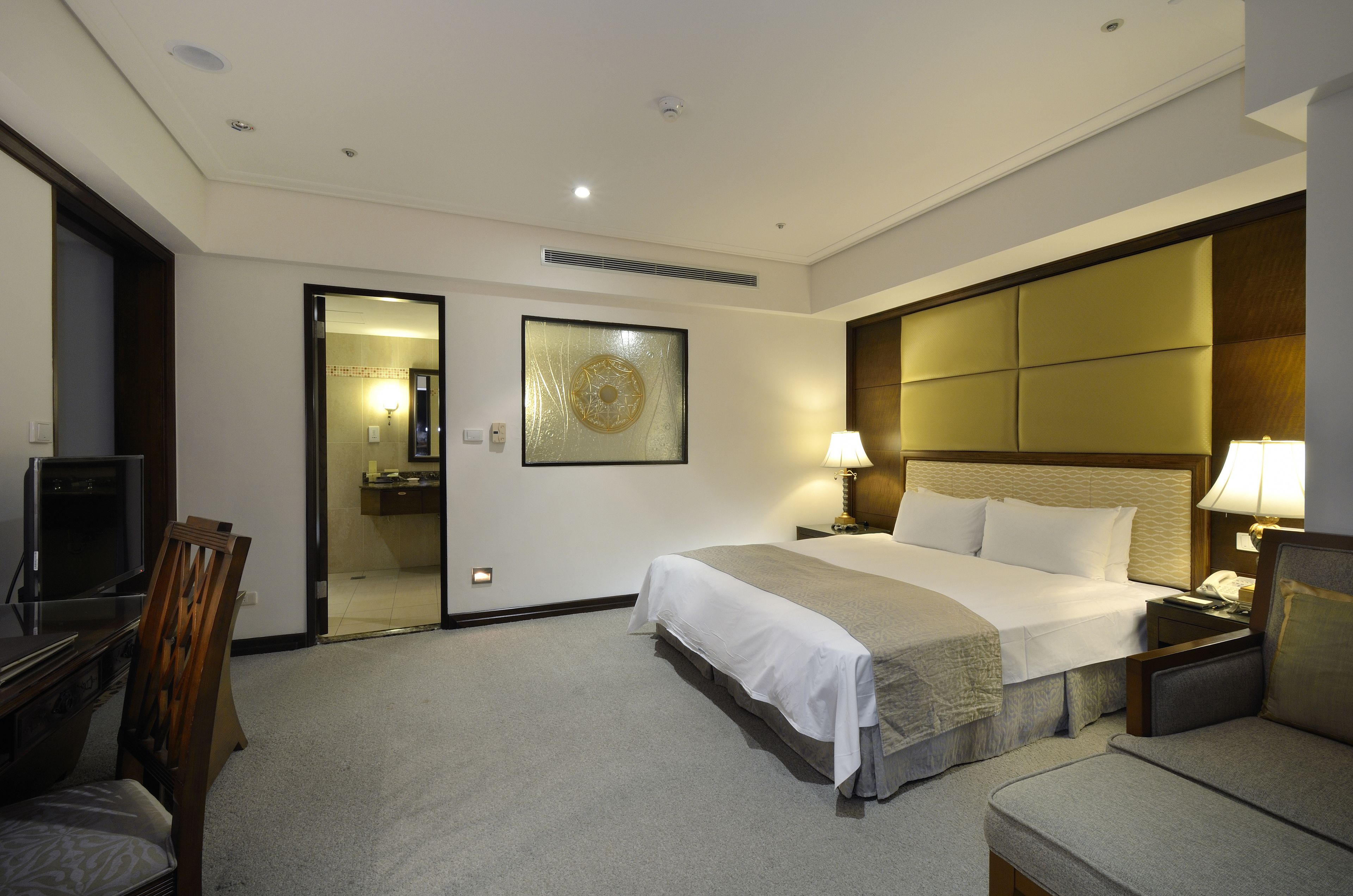A hotel room with a bed, bedside tables, a TV, a chair, and a bathroom visible through an open door.