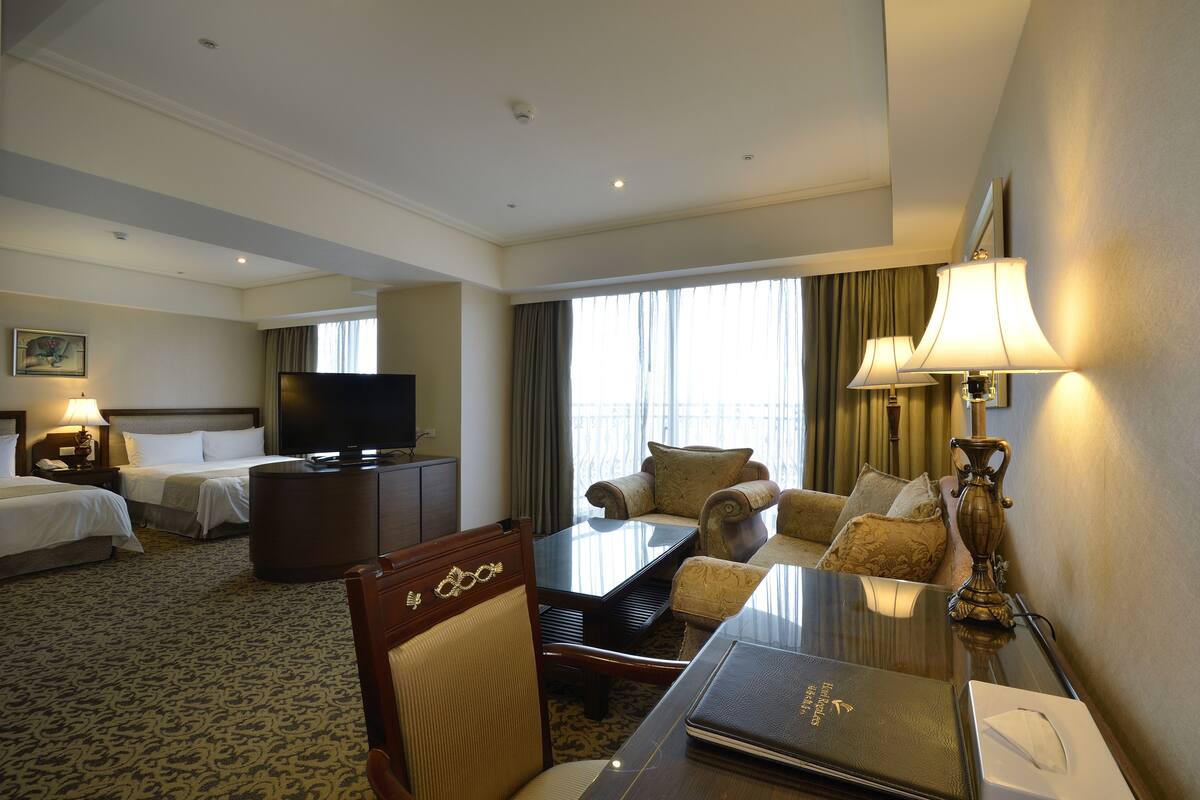 A hotel room with two beds, a TV, a desk with a lamp, and a view of the outside.