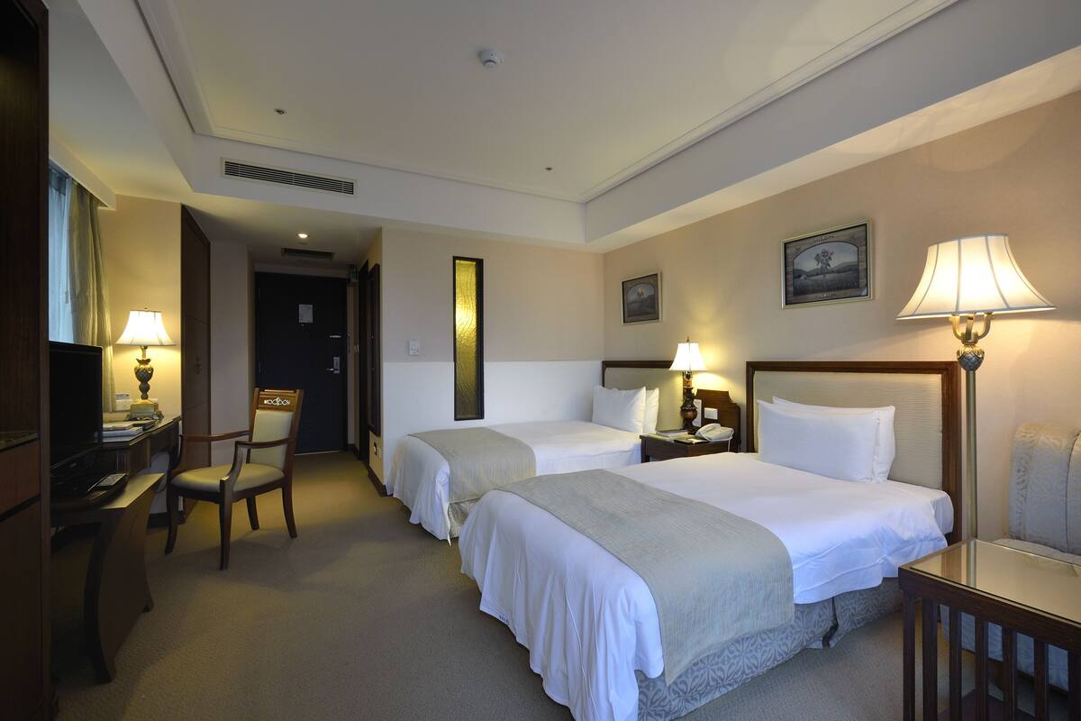 A hotel room with two beds, a desk with a lamp, a chair, and a television.