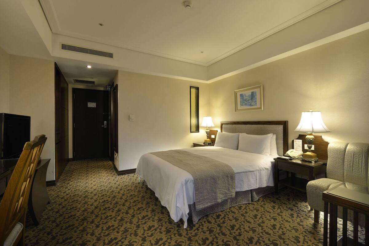 A hotel room with a bed, bedside lamps, a desk, a chair, a television, and a door leading to another room.