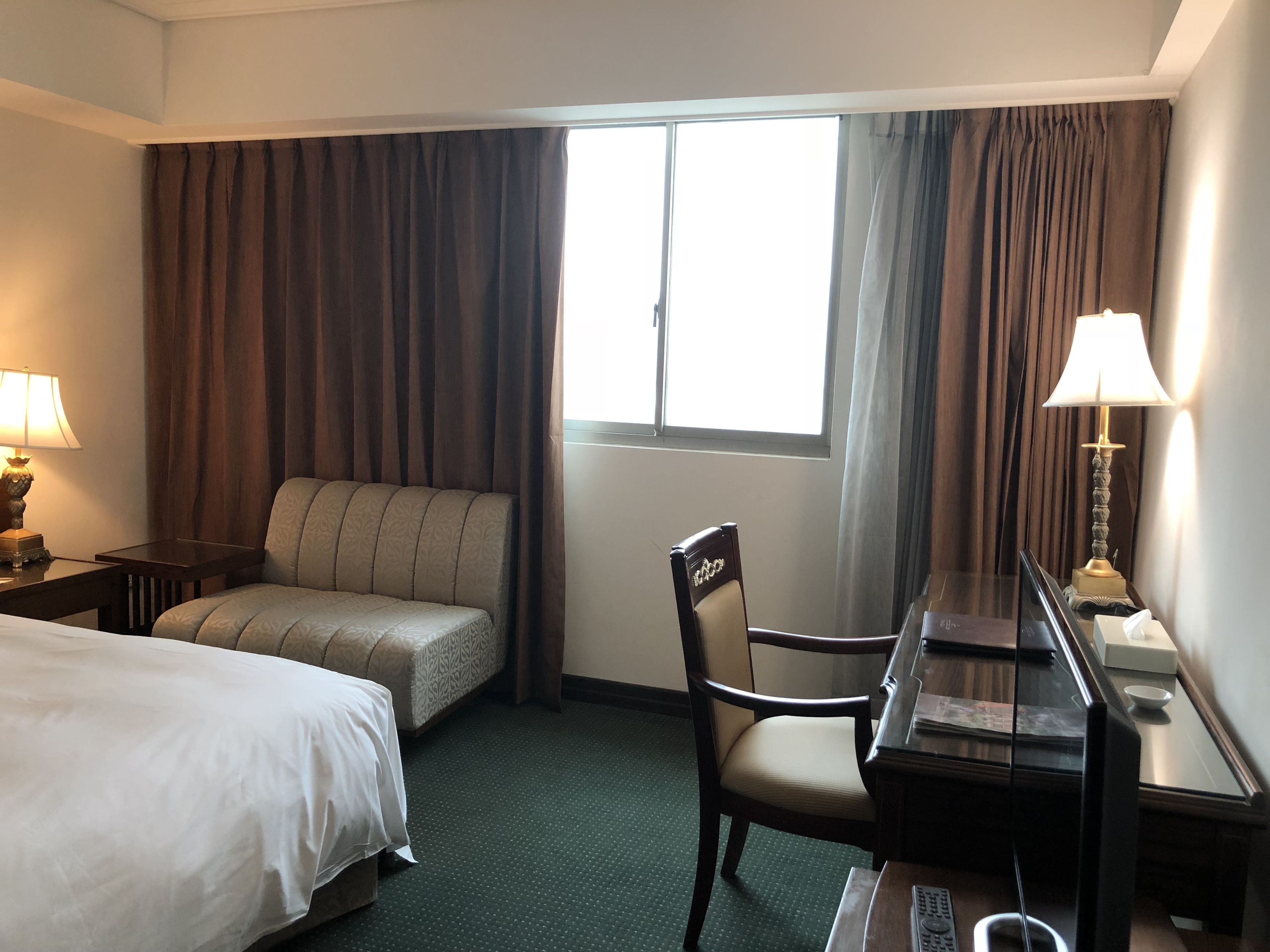 Une chambre d’hôtel avec un lit, un canapé, un bureau et une chaise. Il y a une fenêtre avec des rideaux, une lampe et une table de chevet.
