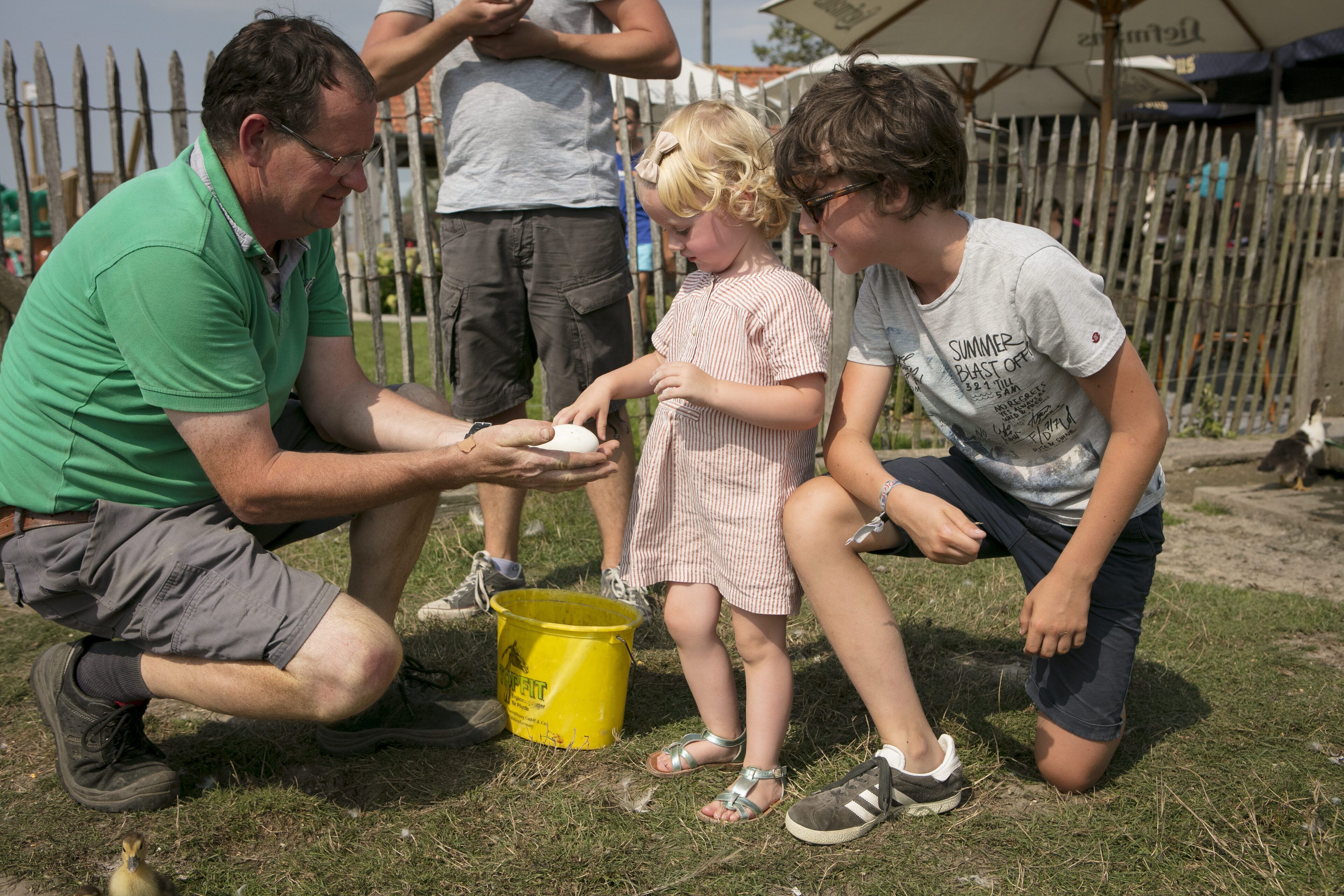 A group of people, including children, gathered around a yellow bucket on a grassy area.