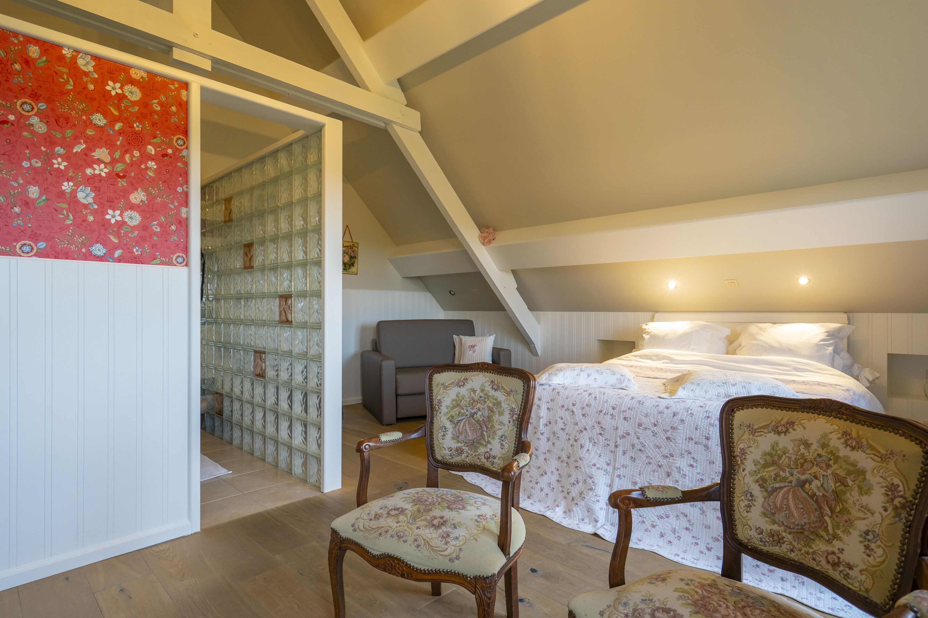 A bedroom with a bed, two upholstered chairs, a small sofa, and a staircase leading to an upper level.