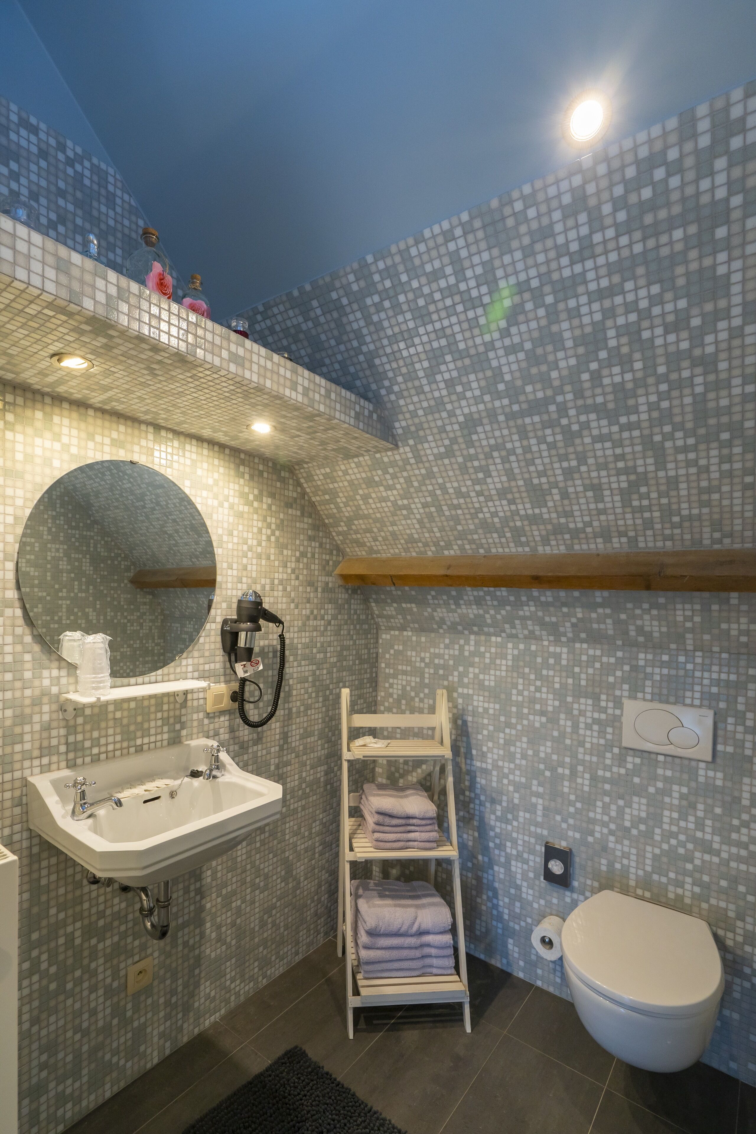 Un baño moderno con una pared de mosaicos, un espejo redondo, un lavabo blanco, un perchero y un inodoro.