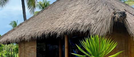Una cabaña con techo de paja, terraza de madera y barandillas de bambú, rodeada de vegetación tropical.