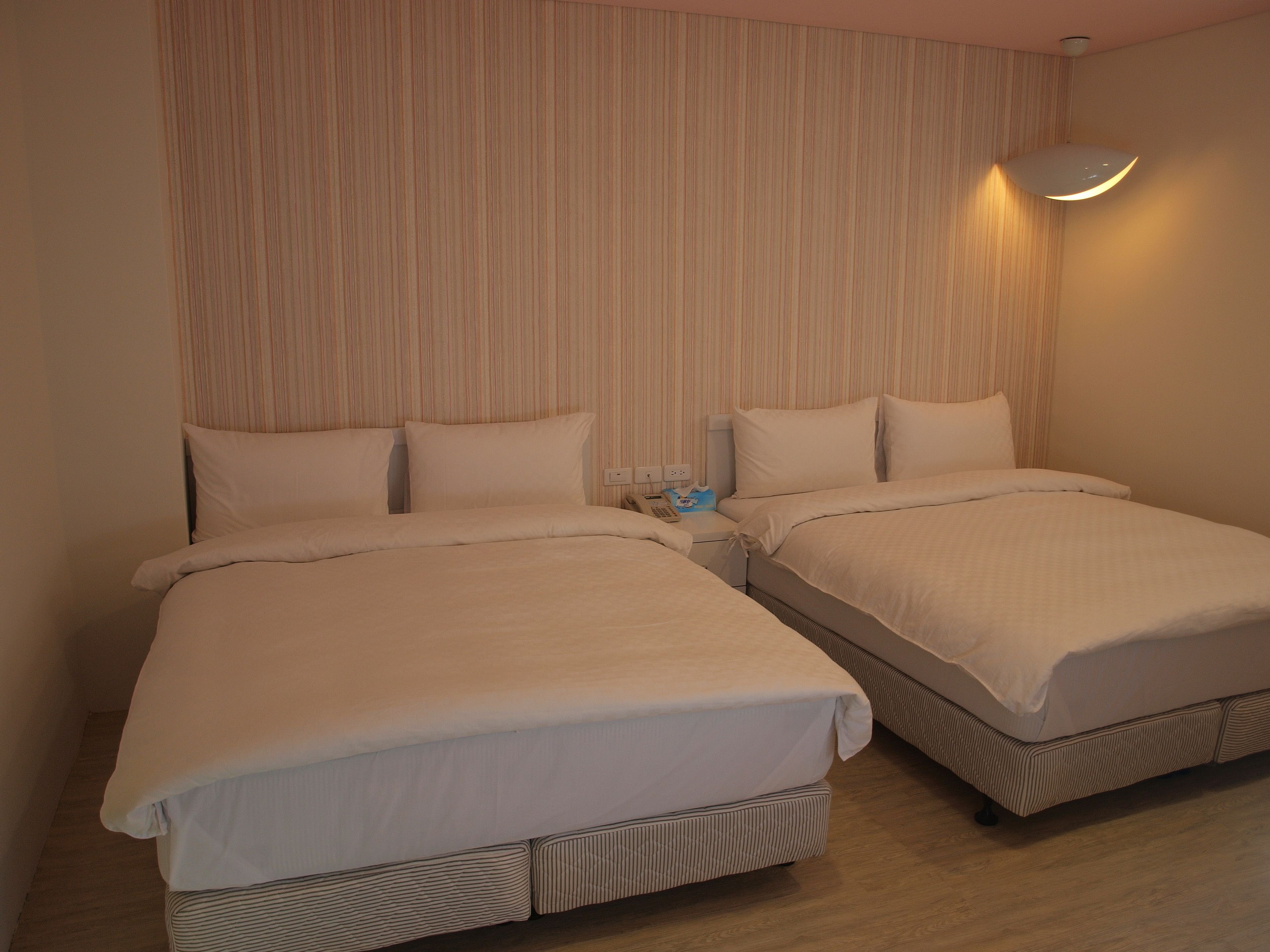A hotel room with two single beds, a bedside table with a phone, and a wall-mounted light fixture.