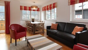Una sala de estar moderna con un sillón de cuero rojo, un sofá negro con cojines estampados, una mesa de centro de madera, y una zona de comedor con una mesa y sillas de madera.