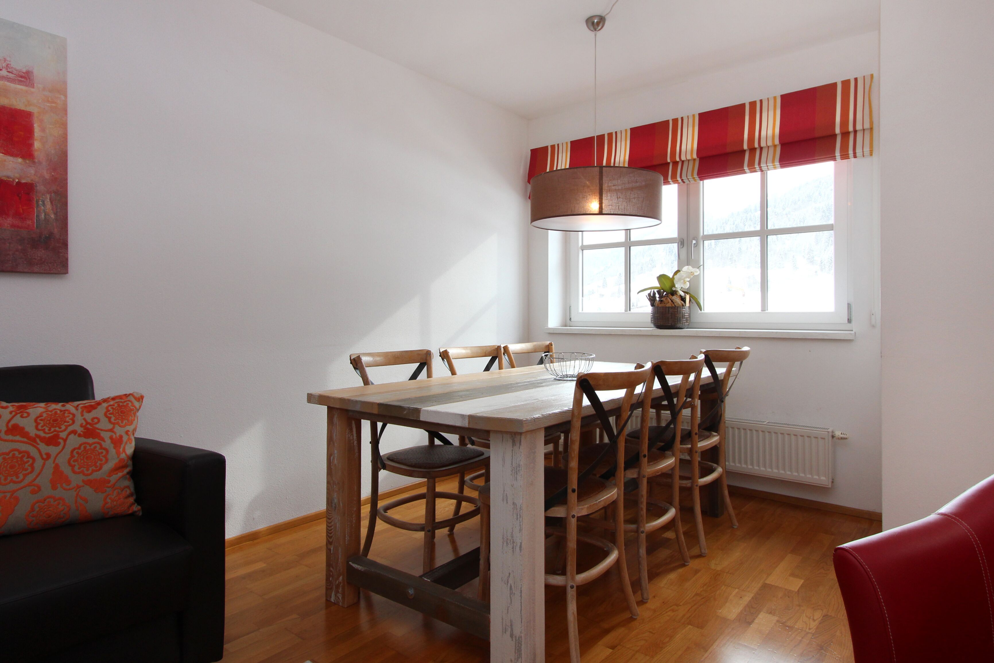 Un comedor con mesa y sillas de madera, un sofá negro y un ventanal con un adorno rojo y blanco.