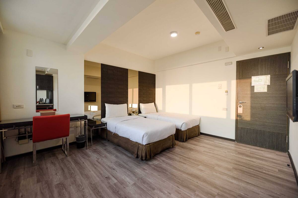 Una habitación de hotel con escritorio, dos camas, una silla roja y un espejo grande.